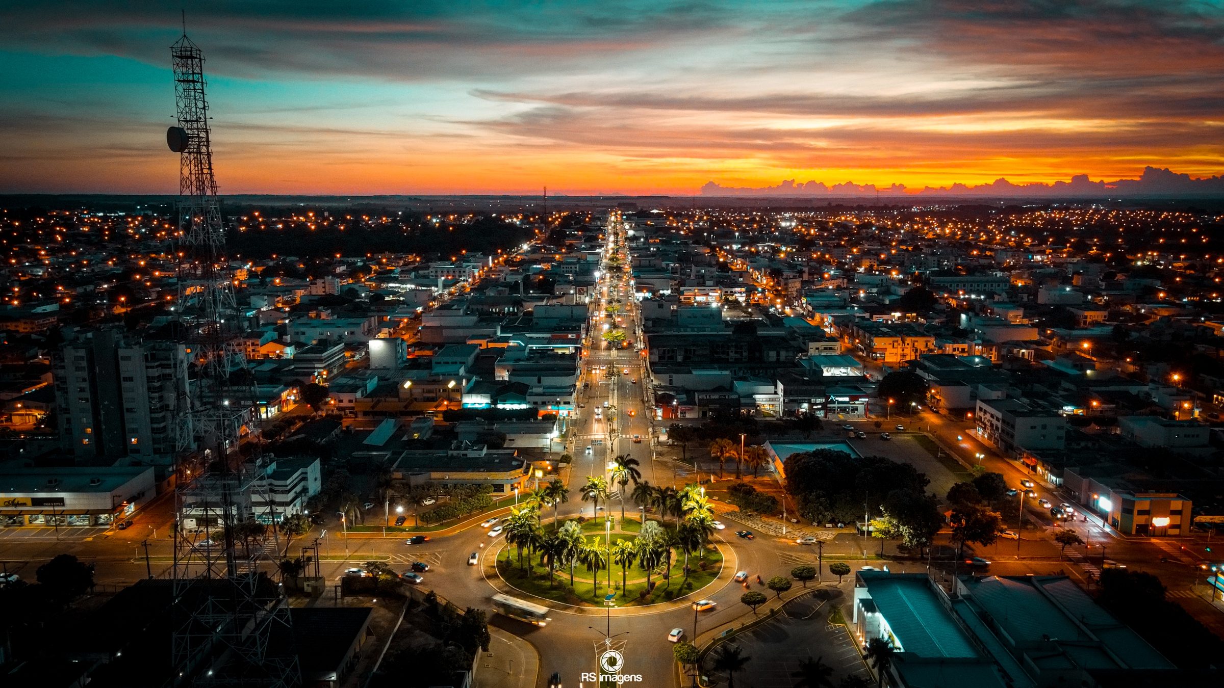 Vista aérea noturna de Tangará da Serra, com a avenida central iluminada e o pôr do sol ao fundo